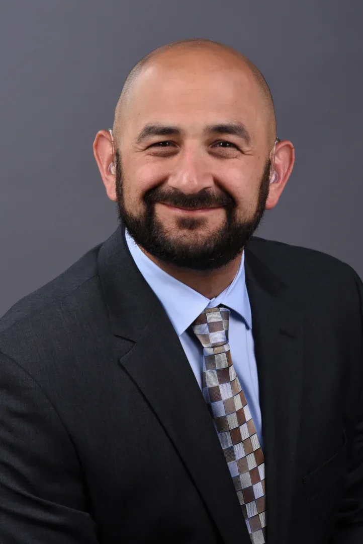 A bald white man with a dark, black beard leans slightly and smiles at the camera. He is wearing a dark suit jacket, collared blue shirt, and a tie with brown, gray, and light blue box patterns.