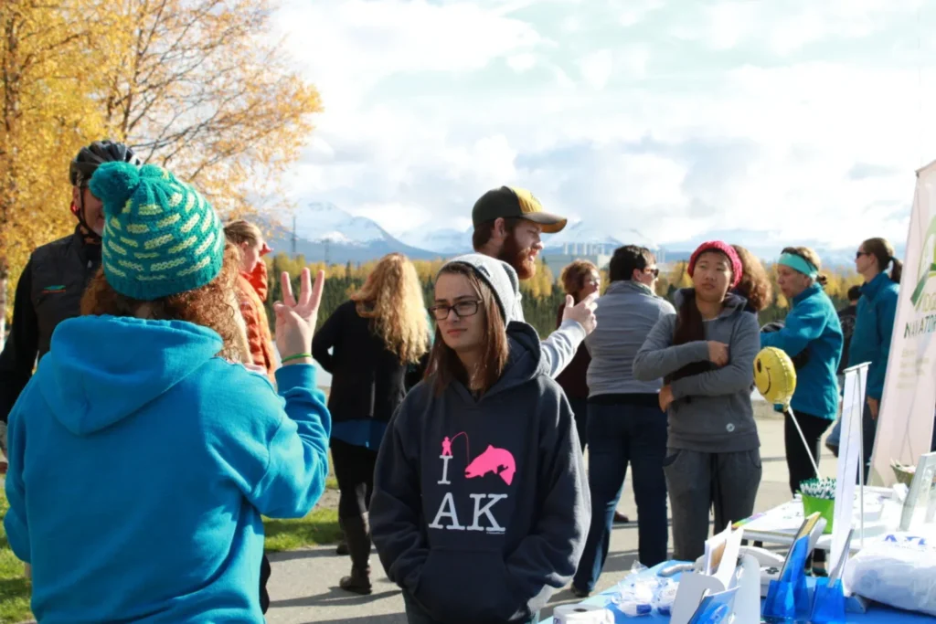 The image is an outdoor scene at a public event with attendees in casual clothing. A person in a grey hoodie with "I ♥ AK" printed on it stands in the foreground. Behind them, various individuals are engaging in conversations. There's an information booth in the background, and the setting includes autumn trees and distant mountains; an Alaskan environment.