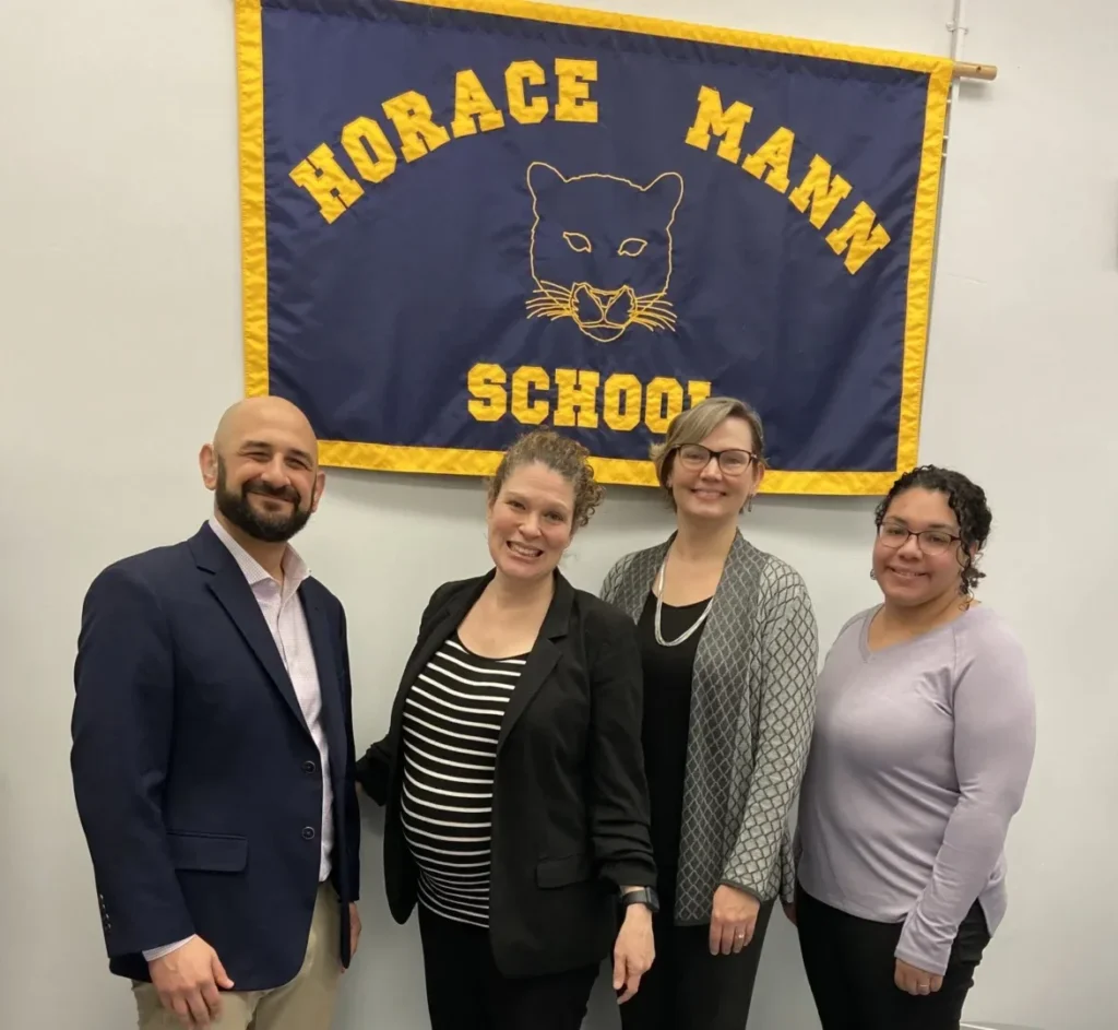 A group picture featuring Shane Feldman and staff at Horace Mann School, in front of the Horace Mann School poster with the partner cat mascot.