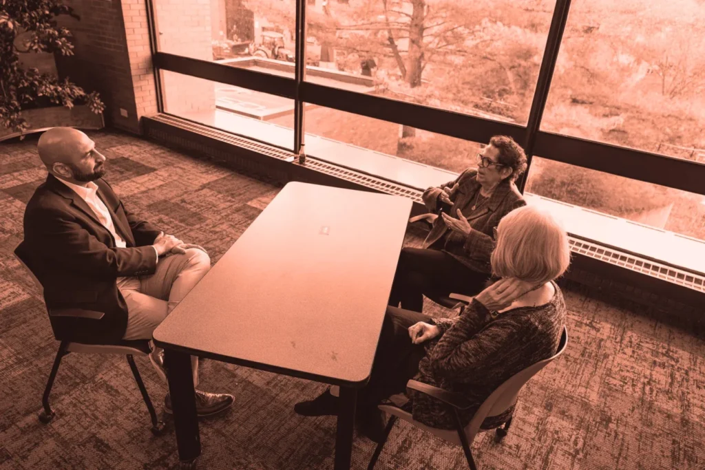 Three individuals are seated around a rectangular table in a room with a large window. One man, wearing a suit, is speaking, gesturing with his hands. Opposite him, an older woman with short hair and glasses listens intently, while a person with short curly hair, wearing a dark jacket, is also engaged in the conversation.