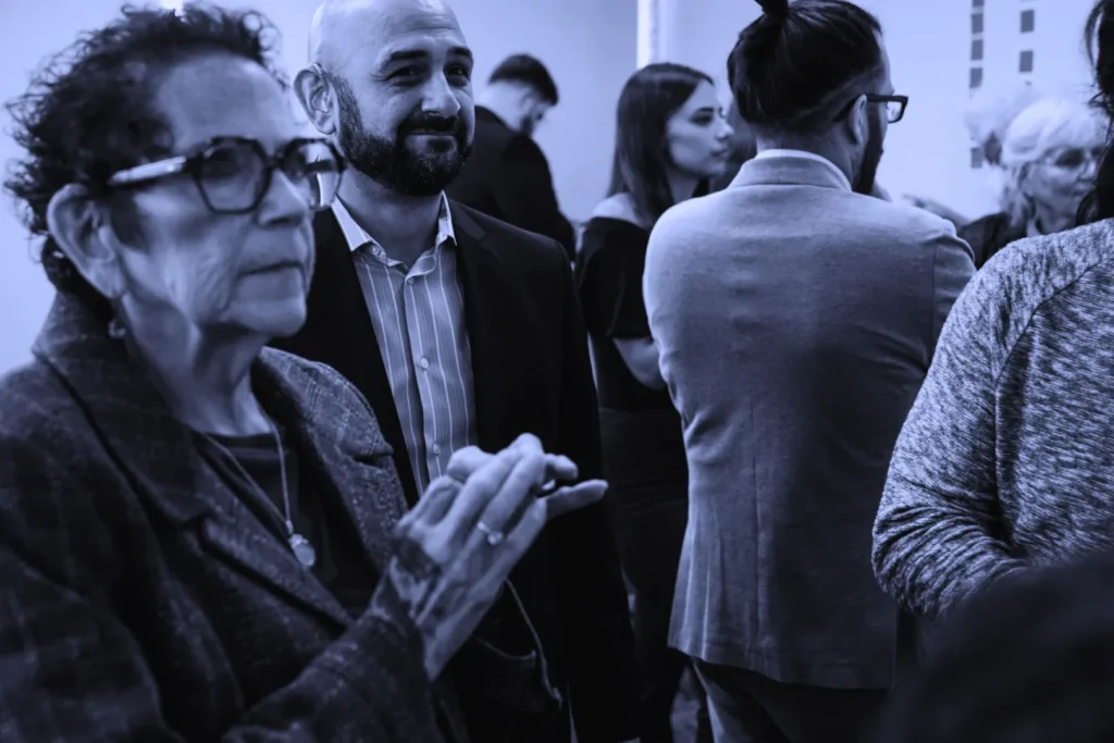 An interactive session with a diverse group of individuals. A woman in a gray blazer is in mid-gesture, speaking to a man with glasses and a man with a man bun, suggesting an exchange of ideas or a discussion.