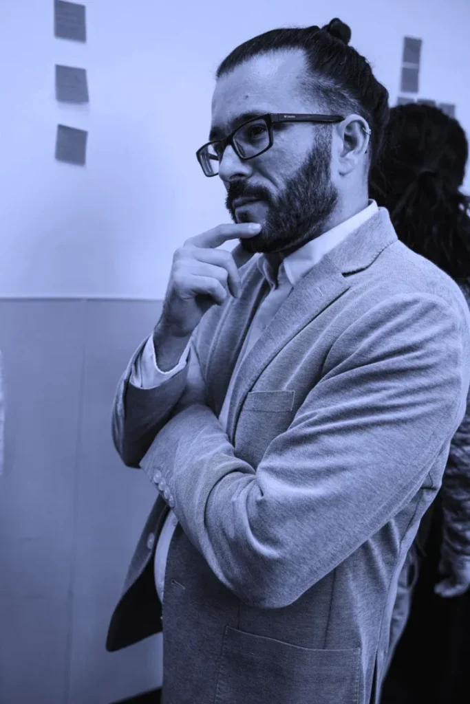 A man with glasses and a bun hairstyle, hand on chin, standing thoughtfully.