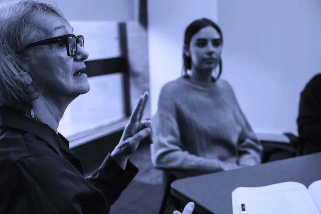 A woman in glasses speaking with her hands raised, with others listening in a classroom setting.