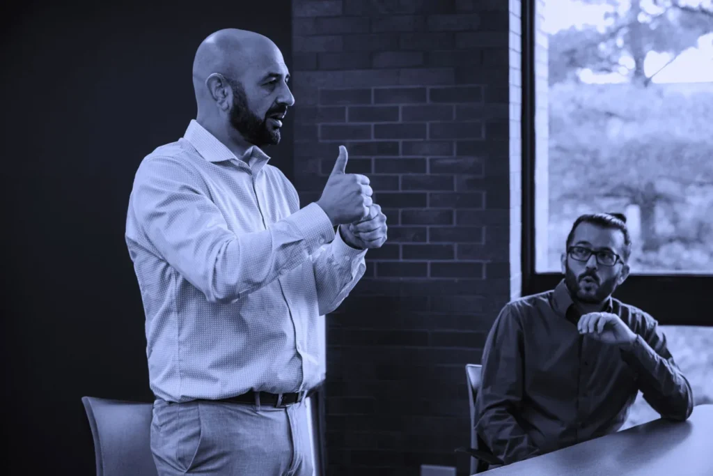 A man standing and giving a thumbs-up gesture while another man sits at a table looking on.