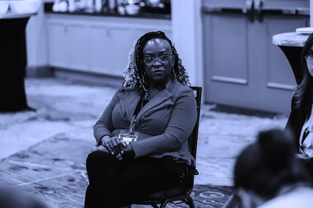 Confident woman in a gray blazer seated and looking attentively towards a speaker out of frame.