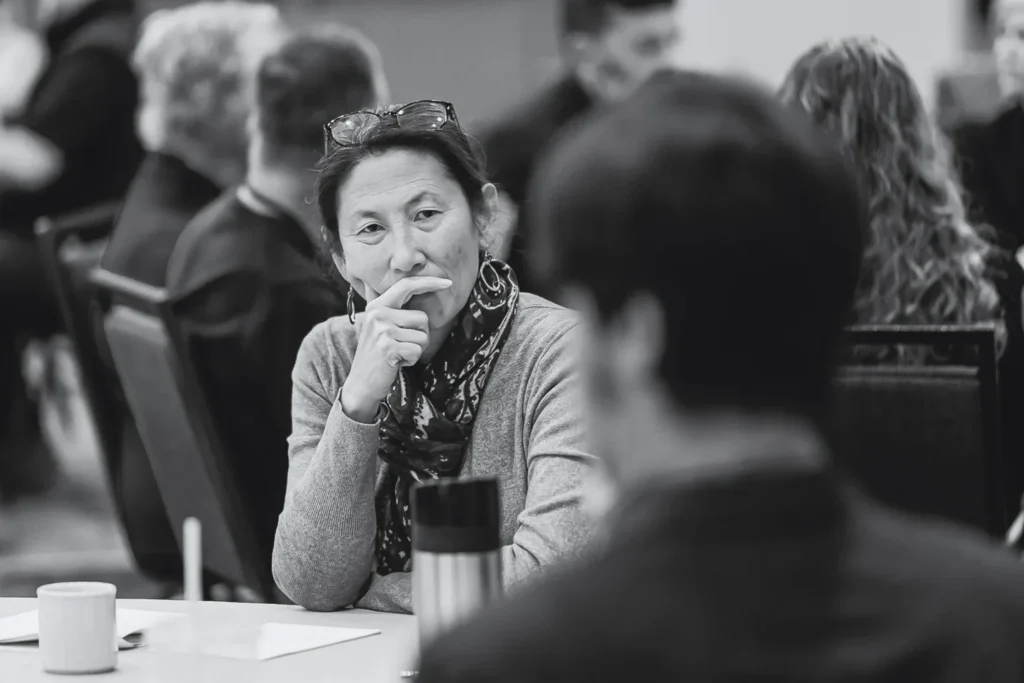 Thoughtful woman touching her chin while listening in a meeting, with other participants in the background.