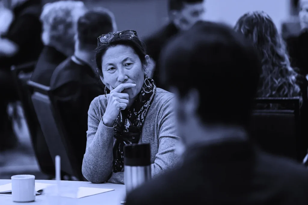 Thoughtful woman touching her chin while listening in a meeting, with other participants in the background.