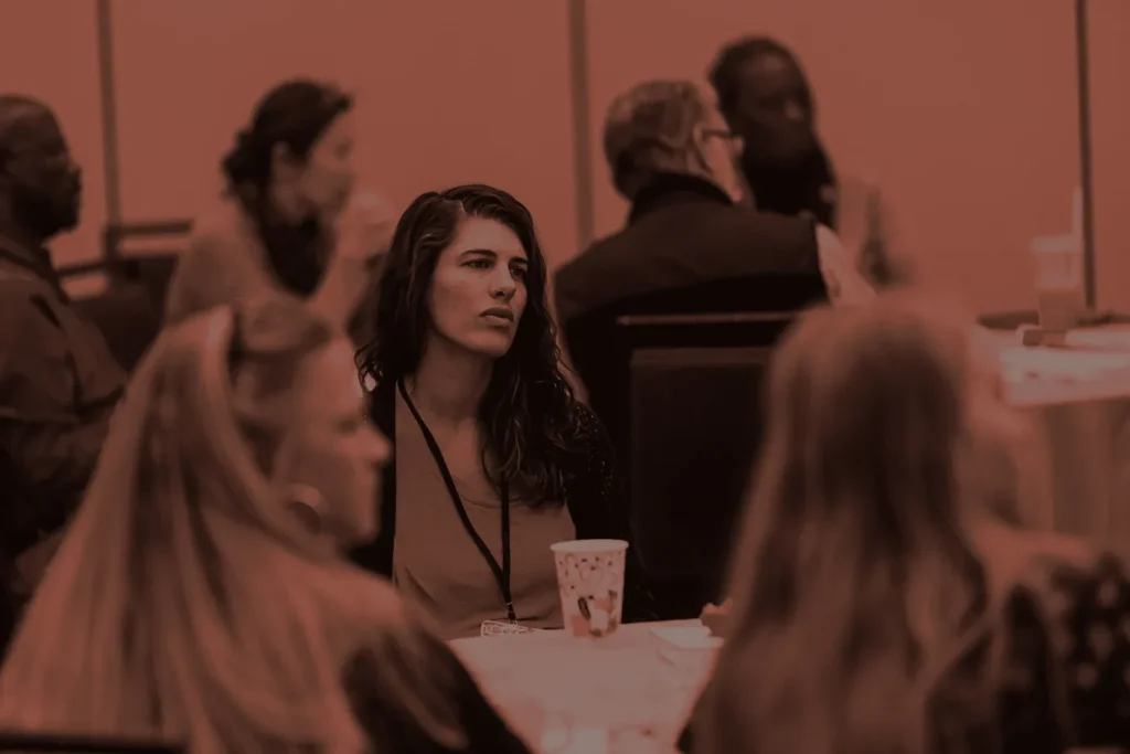 A woman looks on thoughtfully during a conference discussion, with other participants around her.