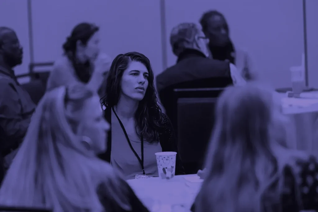 A woman looks on thoughtfully during a conference discussion, with other participants around her.