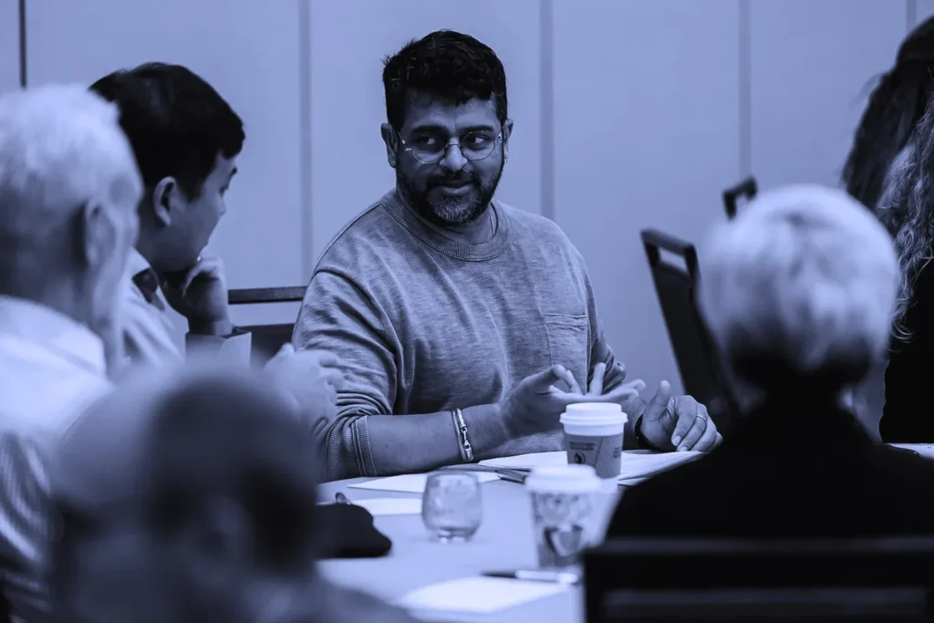 A man with a beard and glasses participates in a group discussion at a conference table.