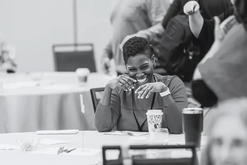 A woman in a red top laughing joyfully with her hands up to her face at a conference table.
