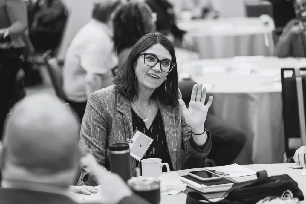 A woman in glasses raising her hand to ask a question in a conference room setting.