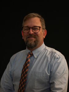 Chris Campbell, a white man with glasses, and a goatee  looks directly ahead with a blue collared shirt and blue and orange pattered tie.