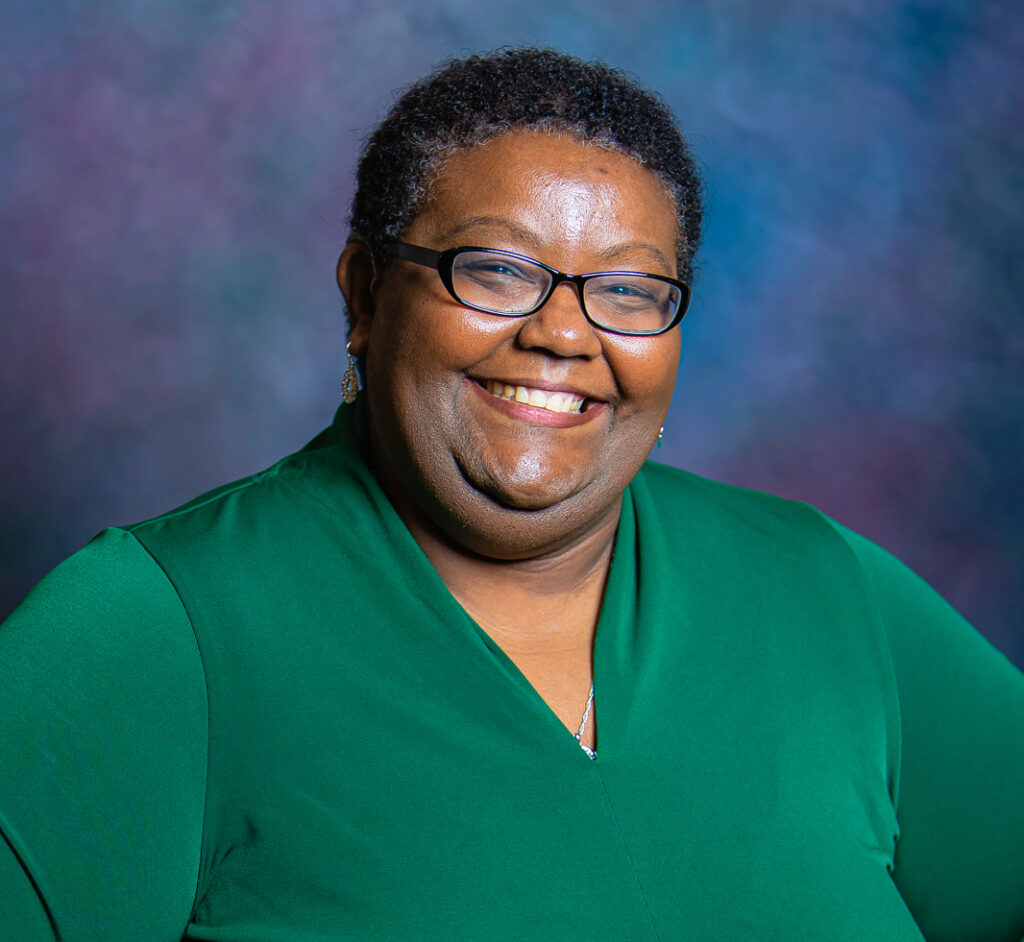 A woman named Darian Gambrell with short curly hair and glasses, wearing a green blouse, smiles at the camera against a blurred multicolored background background.