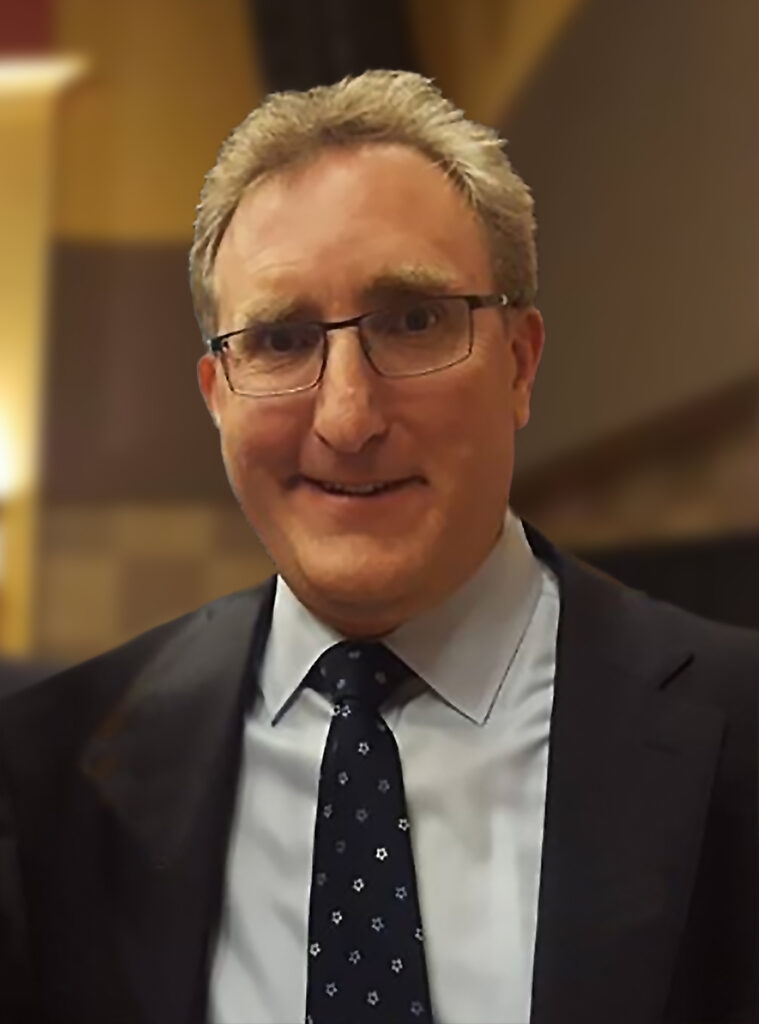 A man with glasses and short hair, wearing a suit and tie, smiling in an indoor setting.