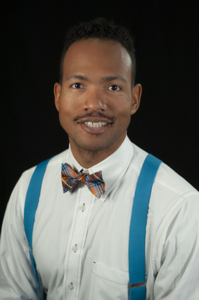 The image is a professional headshot of a man, DJ Johnson against a solid black background. He is wearing a white dress shirt with bright blue suspenders and a colorful plaid bow tie in shades of orange, blue, and brown. He has a short, neatly trimmed mustache and curly black hair. The man is smiling softly and looking directly at the camera, giving a warm and approachable appearance.