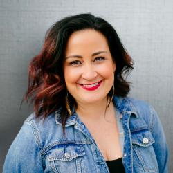 A professional headshot of a woman, Melissa Elmira Yingst with wavy black and red-tinted hair, wearing a denim jacket and a black top. She is smiling against a light gray textured background.