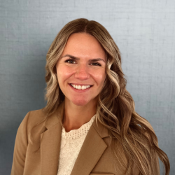 A professional headshot of a woman, Samantha Wulz with long brown/blonde hair, wearing a purple blouse and black blazer. She is smiling and standing against a light gray textured background.