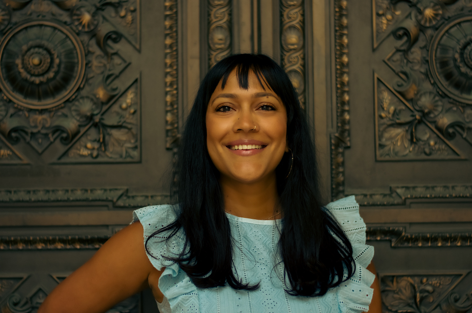 Sasha, a brown woman with long, wavy brown hair, is wearing a light blue dress while standing in front of a decorative iron door.