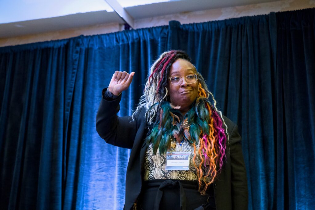 A speaker with multi-colored braids in shades of red, pink, green, and blonde stands in front of a blue curtain, raising one arm slightly. They wear glasses and a name badge, exuding a calm, thoughtful presence on stage