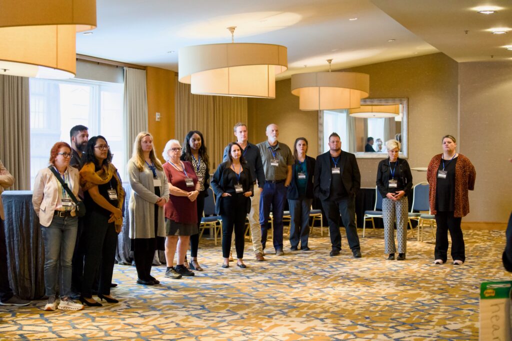 A semi-circle of conference attendees listens attentively to a speaker out of the frame. The room has large light fixtures and windows. Participants wear name badges and business casual attire, creating a collaborative atmosphere