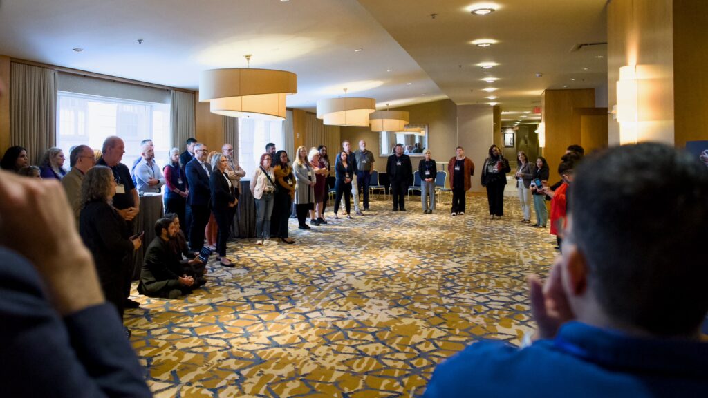 Conference participants form a large circle in a hotel hallway for a discussion or activity. The space has warm lighting, patterned carpet, and large overhead lights. People of various backgrounds face each other, creating a sense of shared focus.