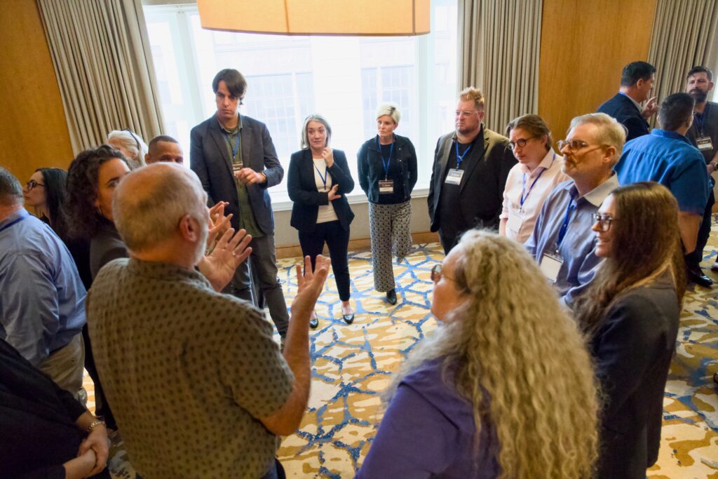 A small group of people stands in a semi-circle in front of a window, listening to a man in a patterned shirt signing in ASL. The setting is a cozy conference room with natural light, patterned carpet, and wooden walls.