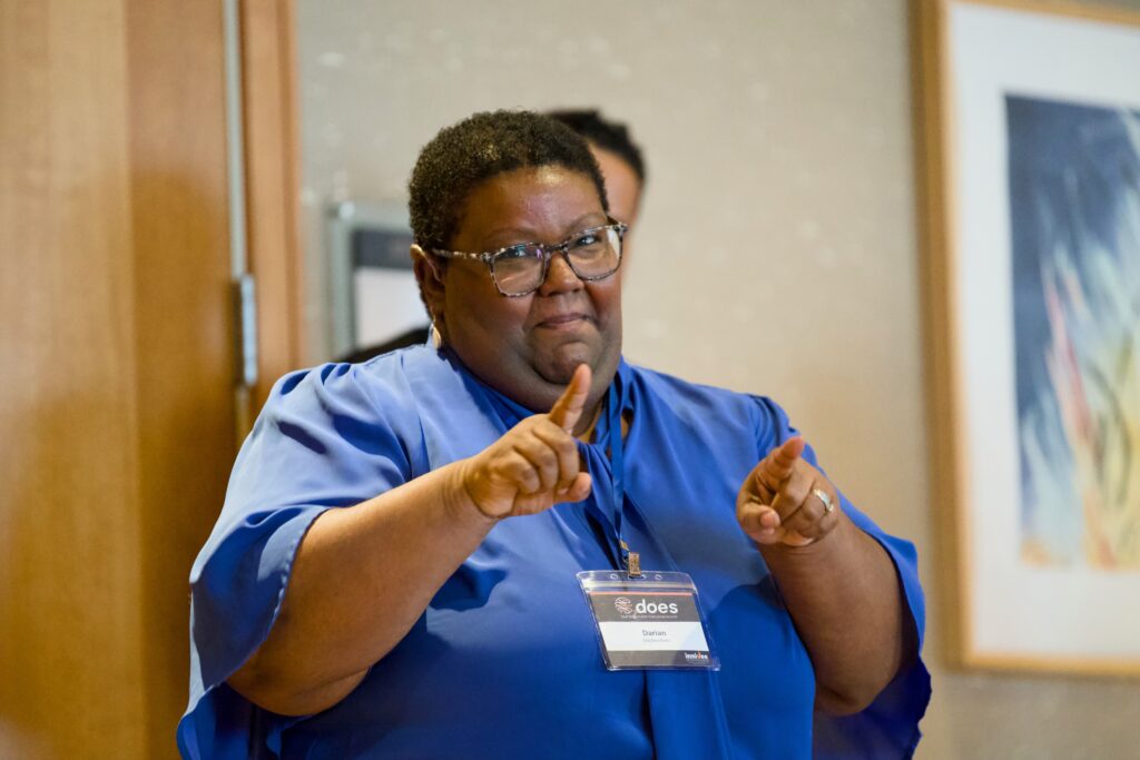 A speaker wearing a royal blue blouse and clear-framed glasses signing in ASL. They stand in front of a neutral-colored wall with artwork, appearing focused and expressive.