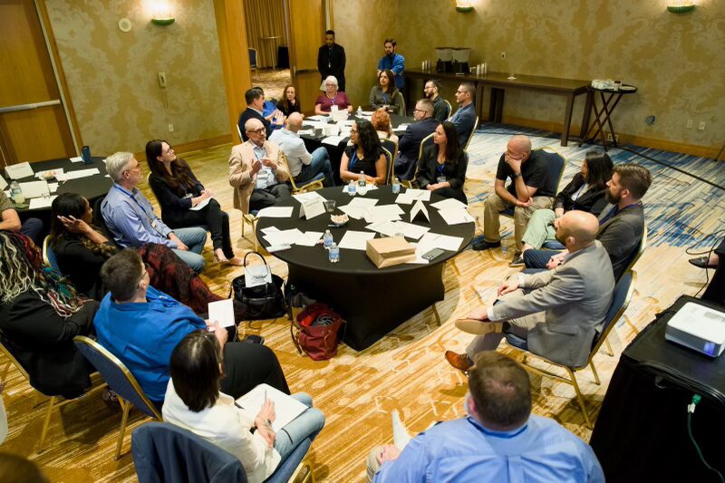 Conference participants form a large circle in a hotel hallway for a discussion or activity. The space has warm lighting, patterned carpet, and large overhead lights. People of various backgrounds face each other, creating a sense of shared focus.