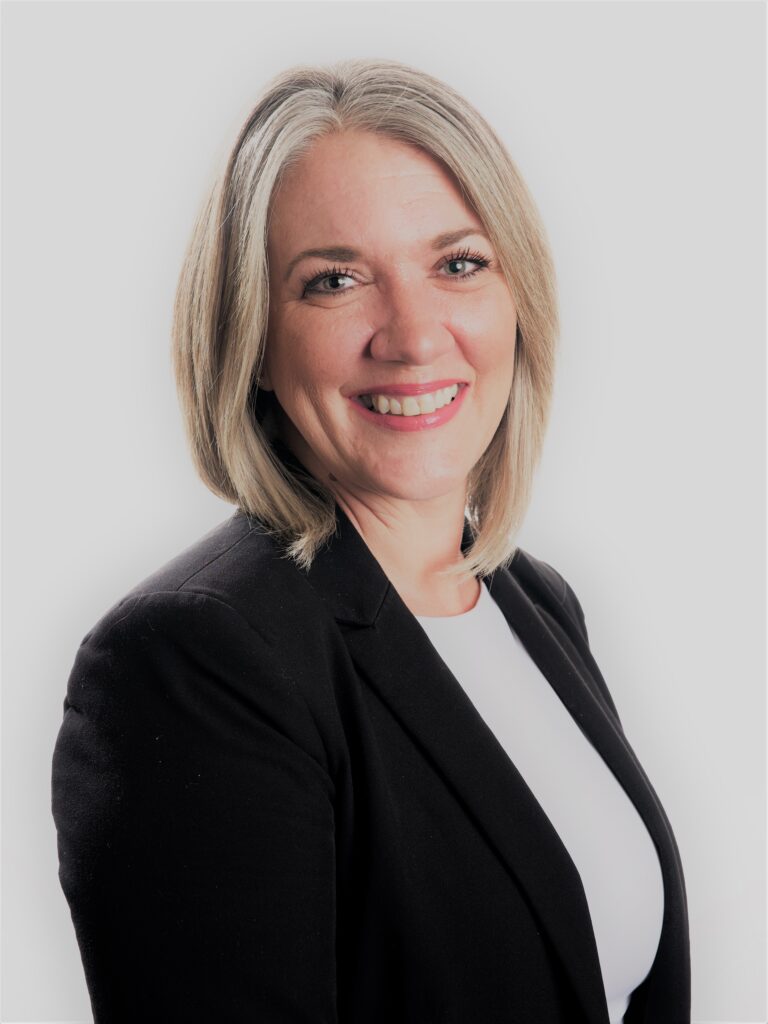 Jana Broussard, a woman with shoulder-length blonde hair smiles warmly at the camera. She is wearing a black blazer over a white shirt, with a plain white background.