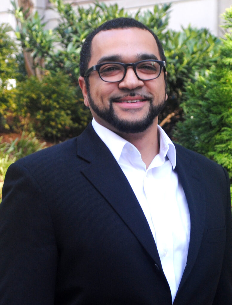 A man, Tim Hopkins in a black suit and white shirt smiles warmly while standing outdoors with greenery in the background.