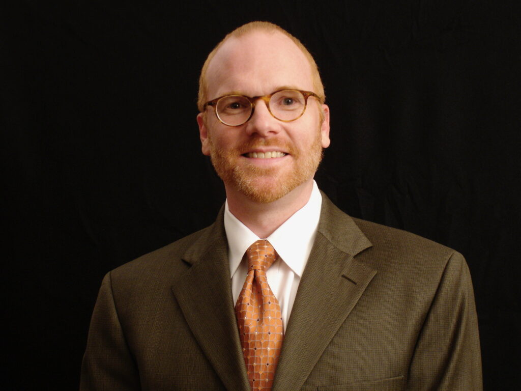 A man, Dr. David Geeslin with short red hair and beard, wearing glasses, a brown suit, white shirt, and orange patterned tie, smiles in front of a black background.