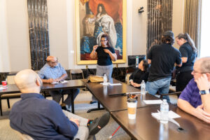 A woman stands in front of a large portrait and signs in ASL during a small group breakout session. Others are seated at tables around her, listening and watching attentively.