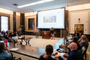 A woman presents in ASL at a podium while a man in a suit stands nearby. The group is watching a projected Facebook post on a large screen behind them.