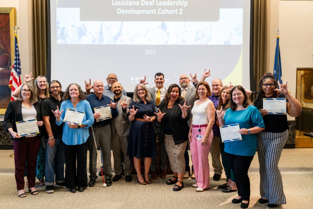 Group photo of LD2 Cohort 2 graduates standing in front of a screen that reads “Louisiana Deaf Leadership Development Cohort 2, 2025.” Many are holding certificates and smiling while signing “I love you” in ASL.