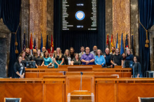 The LD2 cohort poses for a group photo inside the Louisiana Senate chamber in front of the official voting board and flags. Everyone is smiling.