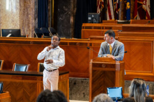 A participant in a white shirt signs in ASL at the Louisiana Senate podium while a suited official watches. Flags and wood-paneled desks frame the scene.