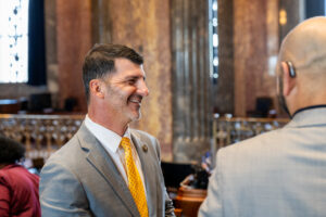 A man in a suit with a yellow tie smiles while shaking hands and chatting with another attendee wearing a hearing device in a formal setting.