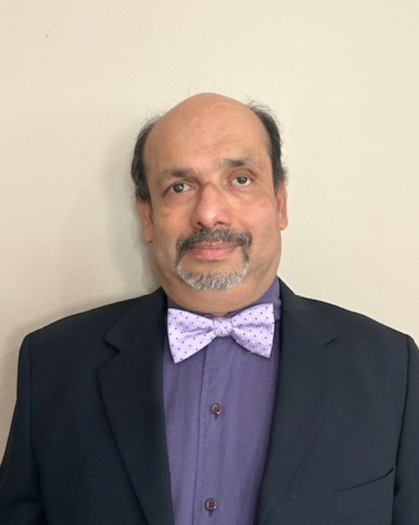 Finalists Announced: Deaf & Hard of Hearing Services (Michigan) Executive Director 2 A man with short dark hair and facial hair, wearing a dark suit jacket, purple shirt, and a light purple bow tie, stands in front of a beige background.