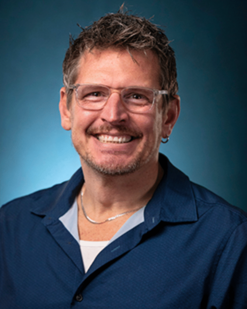 Finalists Announced: Deaf & Hard of Hearing Services (Michigan) Executive Director 1 A man with short brown hair, light facial hair, and glasses, wearing a blue shirt and silver necklace, smiles in front of a blue background.