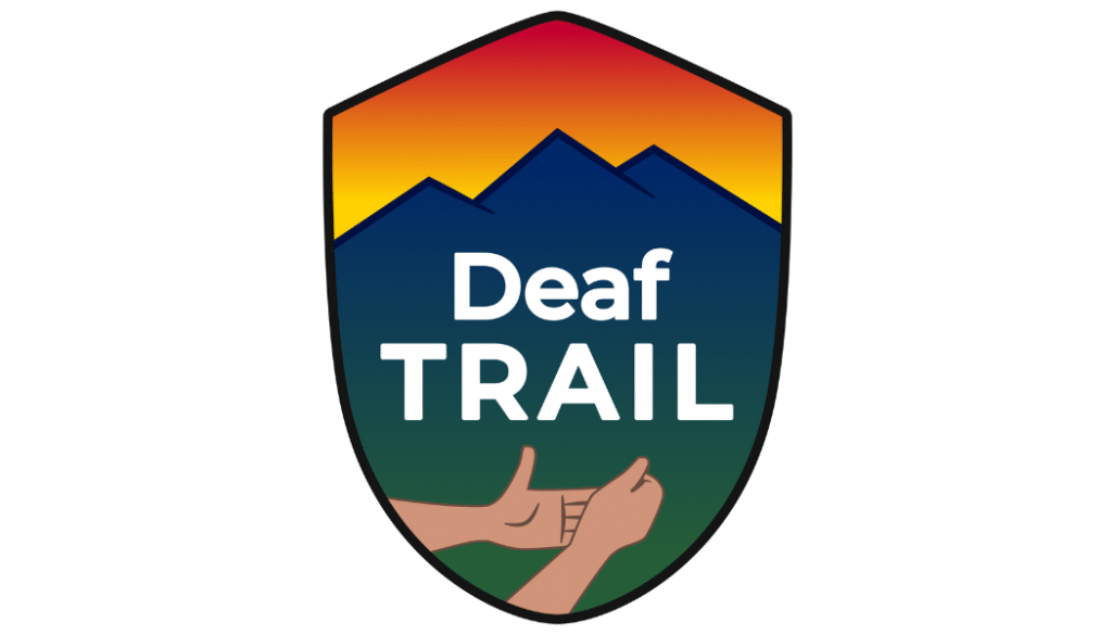 A shield-shaped logo with a mountain silhouette and sunset gradient background. The words “Deaf TRAIL” appear in bold white text, with two illustrated hands below signing “trail” in American Sign Language.
