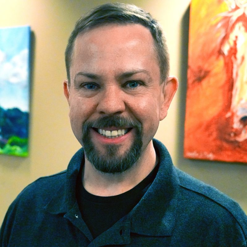 A smiling adult with short light-brown hair and a trimmed beard stands indoors, wearing a dark collared shirt. Colorful artwork hangs on the wall behind them, including a blue-toned landscape and an orange painting featuring part of a horse.