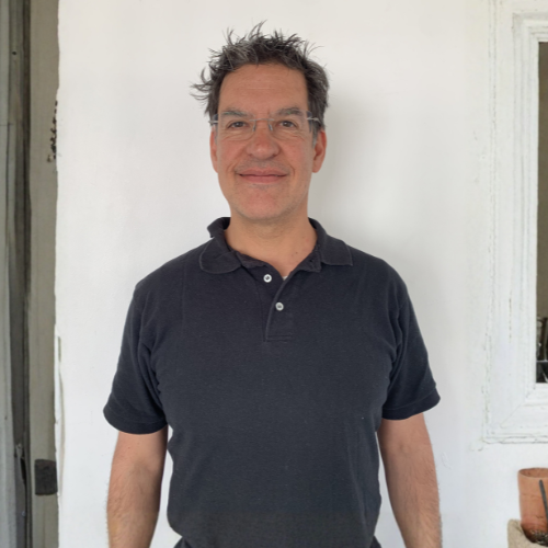 A man with short dark hair and glasses stands smiling in a doorway, wearing a navy polo shirt. He is positioned against a white wall with a small square window to his right. A woven basket with potted plants sits beside him, and green leaves from a nearby plant are visible on the left side of the frame.